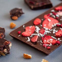 Eine ganze Tafel Schokolade mit eingearbeiteten Erdbeeren und Kokosraspeln. Um die Tafel herum liegen zerbrochene Mandelstückchen und Teile einer anderen zerbrochenen Tafel der gleichen Sorte.