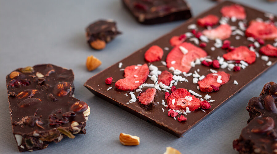 Eine ganze Tafel Schokolade mit eingearbeiteten Erdbeeren und Kokosraspeln. Um die Tafel herum liegen zerbrochene Mandelstückchen und Teile einer anderen zerbrochenen Tafel der gleichen Sorte.